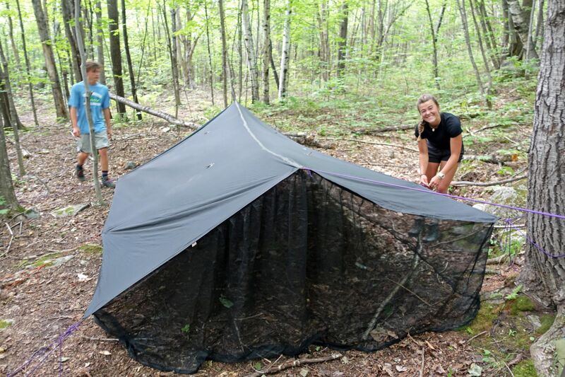 Two people are setting up a dark-colored tent in a forest. The tent has a mesh-like inner layer and is surrounded by trees and foliage. One person is standing near the entrance of the tent, while the other is further back, possibly adjusting the tent's position or securing it. The ground is covered with leaves and small branches.